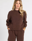 A woman modeling a matching brown knit sweater and shorts set against a plain light gray studio background.