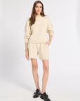 A woman modeling a cream-colored knit sweater and matching shorts set, paired with black leather shoes, against a plain light gray studio background.