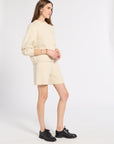 A woman modeling a cream-colored knit sweater and matching shorts set, paired with black leather shoes, against a plain white studio background.