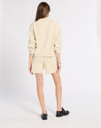 A model is shown from the back wearing a cream-colored knit mock-neck sweater and matching shorts, paired with black leather shoes, against a plain white studio background.