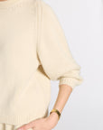 A close-up of a person wearing a cream-colored ribbed knit sweater with a gold watch on their wrist, set against a plain light gray background.