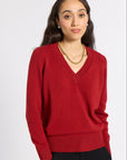A woman modeling a red V-neck knit sweater with black trousers, standing against a plain light gray studio background.