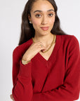 A woman wearing a red V-neck knit sweater, styled with a gold chain necklace, gold watch, and gold ring, against a light gray studio background.