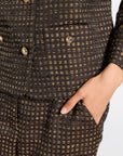 Close-up of a textured black and gold tweed jacket with gold buttons, featuring a hand tucked into a side pocket.