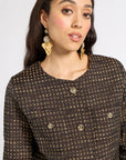 A close-up of a woman wearing a dark brown tweed blazer with gold buttons and matching gold statement earrings against a light gray background.