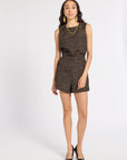 A woman modeling a sleeveless black romper with a textured gold geometric pattern, paired with gold jewelry and black heels, against a plain white studio background.