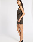 A woman modeling a sleeveless black mini dress with a textured gold geometric pattern, paired with black pointed-toe heels, photographed against a plain white studio background.