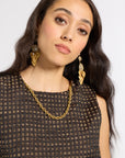 A woman with long dark wavy hair wearing a textured black top with gold lettering, a gold chain necklace, and large gold leaf-shaped drop earrings, photographed against a light gray studio background.