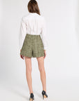 A model shown from the back wearing a white long-sleeve button-up shirt, olive green and beige plaid shorts, and black high heels with gold heels, against a plain white studio background.