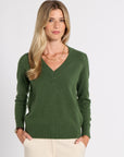 A woman modeling a dark green v-neck knit sweater with cream pants, standing against a light gray studio background.