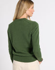 A woman shown from the back wearing a dark green knit sweater and cream-colored trousers, photographed against a clean white studio background.