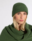 A woman wearing a ribbed knit beanie and matching cowl neck scarf in olive green, photographed against a plain light gray studio background.