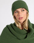 A woman wearing a ribbed knit beanie and matching cowl neck scarf in olive green, photographed against a plain light gray studio background.