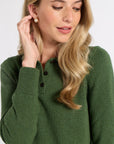 A woman with blonde hair wearing a green knit henley sweater and a small pearl stud earring, shown in a close-up shot against a light gray background.