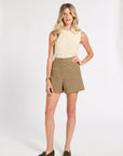 A woman modeling a sleeveless cream cable-knit top paired with brown and beige plaid shorts, styled with dark olive green loafers against a plain white studio background.