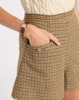Close-up of a person wearing high-waisted plaid shorts in a tan, brown, and black plaid pattern, paired with a cream cable-knit top. The person's hand is in the pocket of the shorts.