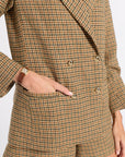 A close-up of a person wearing a brown and beige houndstooth blazer and matching shorts, with one hand in a pocket and a gold watch visible on the wrist.