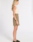 A woman stands in profile wearing a cream sleeveless knit top and a brown and beige houndstooth mini skirt with dark olive loafers, photographed against a seamless white studio background.