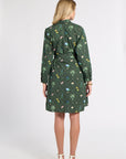 A woman viewed from the back wearing a dark green long-sleeve shirt dress with a colorful floral and botanical print, paired with white high-heeled sandals, against a plain light gray studio background.