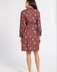 A woman is shown from the back wearing a long-sleeved, knee-length dress with a multicolor floral and botanical pattern on a dusty rose base, paired with beige flat shoes against a white studio background.