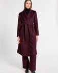 A woman modeling a long burgundy wrap coat with a matching waist tie, paired with matching wide-leg trousers, a black turtleneck, and black pointed-toe heels, against a plain light gray studio background.