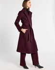 A woman modeling a long, belted cashmere coat in deep burgundy, paired with matching trousers and black pointed-toe heels, photographed against a plain light gray studio background.