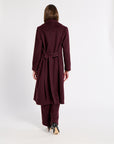 A woman viewed from behind wearing a long burgundy cashmere belted coat and matching trousers against a plain white studio background.