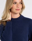 A woman with blonde hair wearing a navy blue ribbed knit sweater, shown in a close-up portrait against a light gray background.