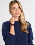 A blonde woman wearing a navy blue knit crewneck sweater, posing with her hand near her neck against a light gray studio background.