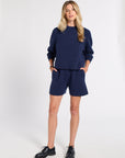 A woman modeling a matching navy blue knit lounge set consisting of a crewneck sweatshirt and shorts, paired with black leather shoes, against a plain light gray studio background.