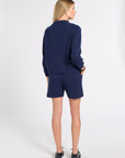 A woman stands with her back to the camera wearing a navy blue knit sweater and matching knit shorts, paired with black shoes, against a plain white studio background.