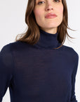 A close-up of a woman wearing a dark navy blue turtleneck sweater against a plain light gray background.
