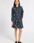 A woman modeling a dark navy blue long-sleeve shirt dress with a colorful floral and botanical print, featuring a button-up front, collar, and self-tie waist belt, paired with dark loafers against a light gray studio background.