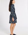 A woman stands in profile wearing a dark navy long-sleeve mini dress with a colorful floral and butterfly print, paired with dark navy loafers, against a plain light gray studio background.