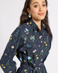 A woman wearing a dark navy long-sleeve button-up shirt with a colorful floral and insect print, featuring a matching waist tie belt, set against a plain light gray background.