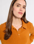 A woman with long brown hair wearing a mustard orange knit polo shirt, photographed against a clean light gray studio background.