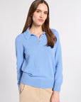A woman wearing a light blue knit polo sweater and beige trousers, standing against a plain light gray studio background.