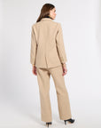 A woman is shown from the back wearing a beige tailored suit consisting of a blazer and wide-leg trousers, paired with black pointed-toe heels, against a plain light gray studio background.