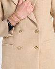 Close-up of a beige double-breasted blazer with gold buttons, worn by a model whose hand and wrist with gold bracelets are visible.