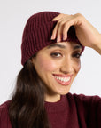 A smiling woman with long dark hair wearing a ribbed knit beanie and matching knit sweater against a plain light gray background.