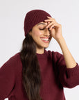 A woman wearing a ribbed burgundy knit beanie and a matching burgundy knit sweater, smiling against a light gray studio background.