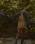 Two people wearing outdoor gear, including the Men's Redford Crew in navy, walking hand in hand through a shallow stream in a forest setting.