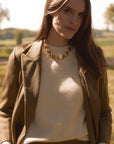 Model wearing the cream Classic Cashmere Crew under a brown blazer, accessorized with a chunky gold necklace, standing outdoors in sunlight.