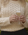 Close-up shot of model holding a glass of champagne in the cream chunky cable knit fisherman crewneck, paired with black slacks.