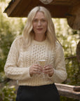 Blonde woman wearing the chunky Aran cable knit fisherman crewneck in cream, holding a glass of champagne in an outdoor, rustic setting.
