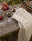 Chunky Aran cable knit fisherman crewneck in cream on a rustic table with apples and a glass of wine, creating a cozy autumn scene