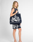A woman wearing a navy black crochet dress and holding a matching black crochet tote bag with white floral embroidery, against a light gray studio background. Styled with black mule pumps.
