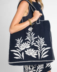 A woman wearing a navy black crochet dress and holding a matching black crochet tote bag with white floral embroidery, against a light gray studio background.