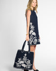 A woman stands in profile wearing a sleeveless black dress with large white floral embroidery and holding a matching black tote bag with white floral embroidery, against a light gray studio background.
