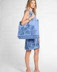 A woman modeling a light blue crochet dress with a dark blue tropical floral print, carrying a matching crochet tote bag, against a light gray studio background.
